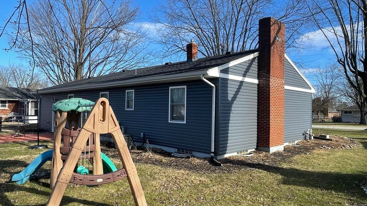 A side view of a blue, single-story house with a brick chimney and a wooden playset in the backyard.