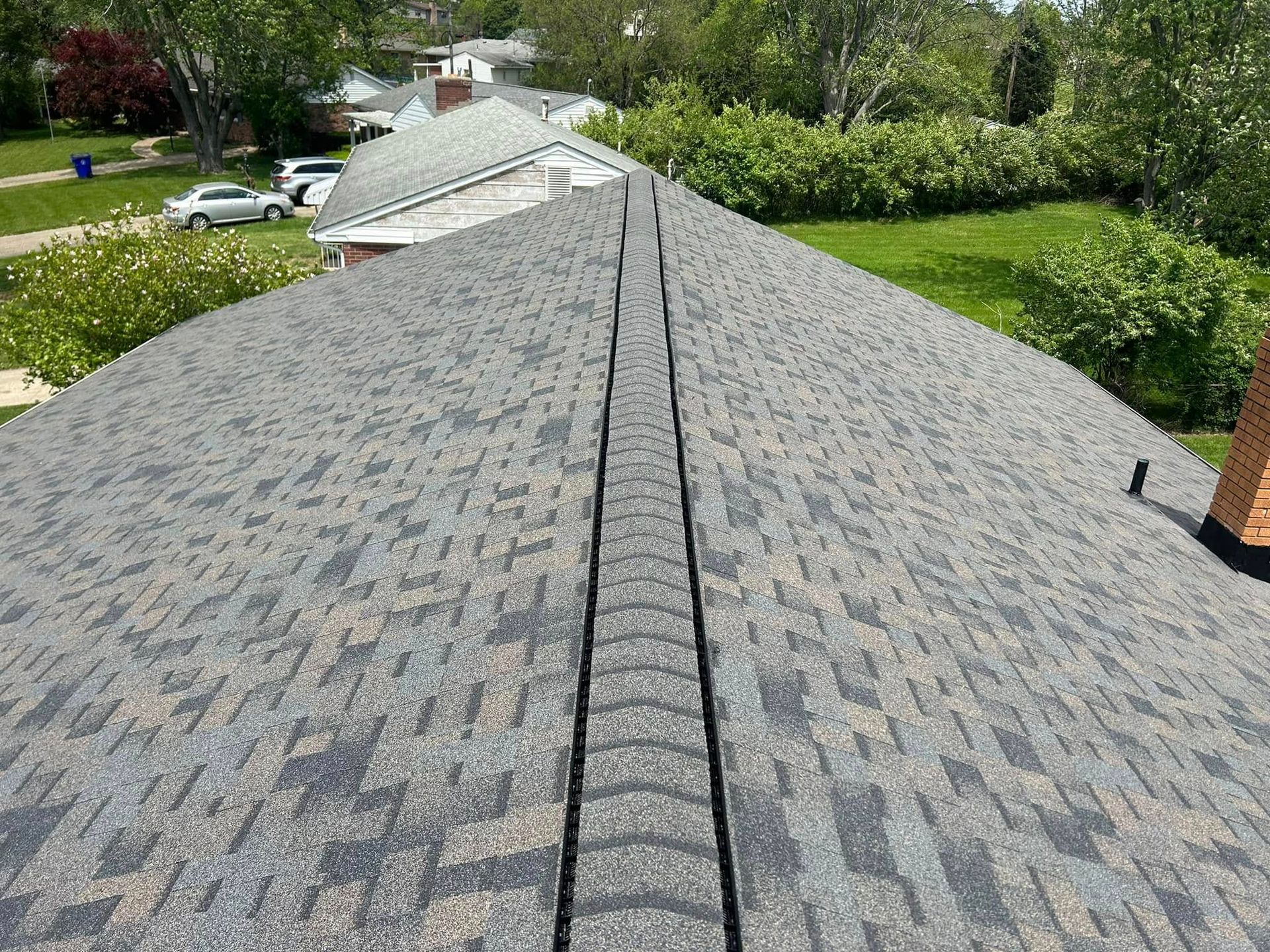 A high-angle view of a gray shingled roof with a central ridge vent, featuring a suburban backyard in the background.