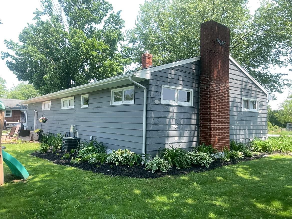 A one-story gray house with a prominent red brick chimney and white window frames, surrounded by garden beds and lawn.