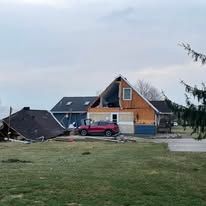 A house with severe storm damage, missing part of its roof and siding, sits next to a red car in a rural landscape.