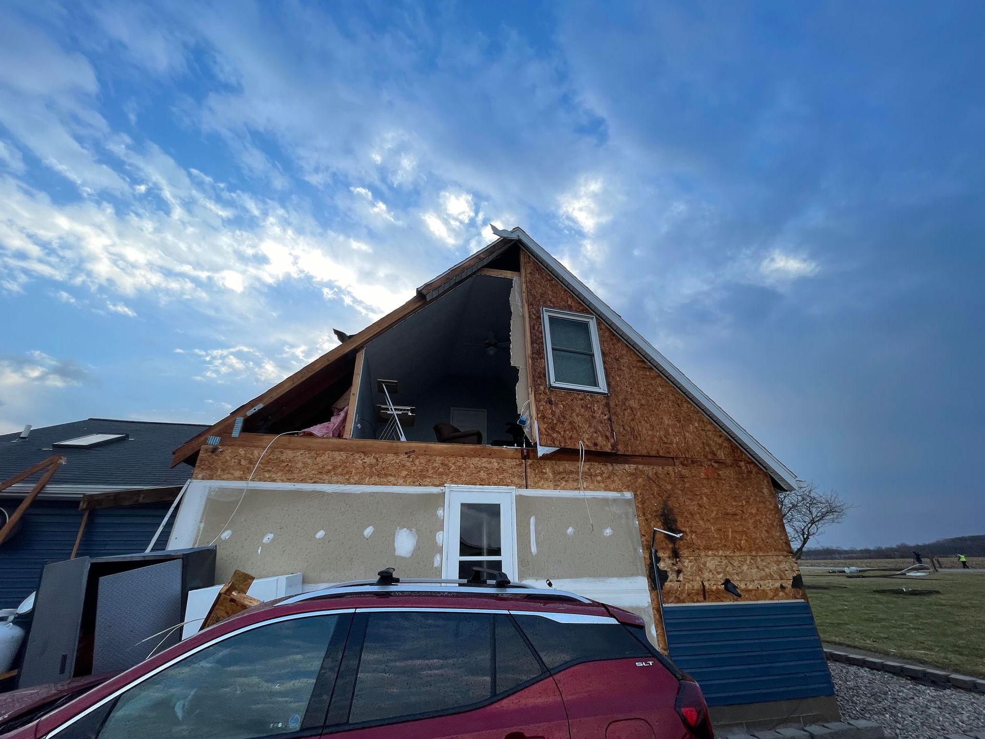 A damaged house with a missing roof and wall, viewed from the side behind a red car under a partly cloudy blue sky.