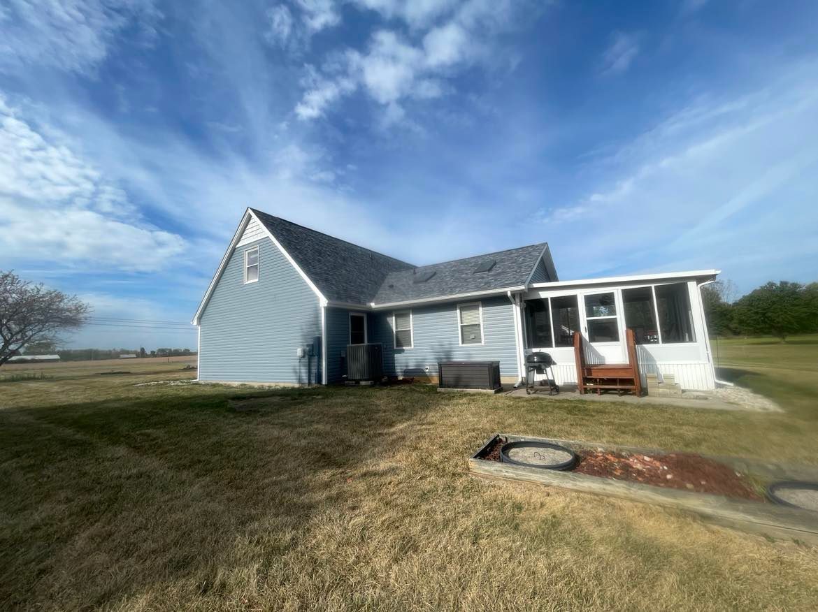 A light blue house with a gray roof and a white screened-in porch sits on a grassy field under a sunny blue sky.