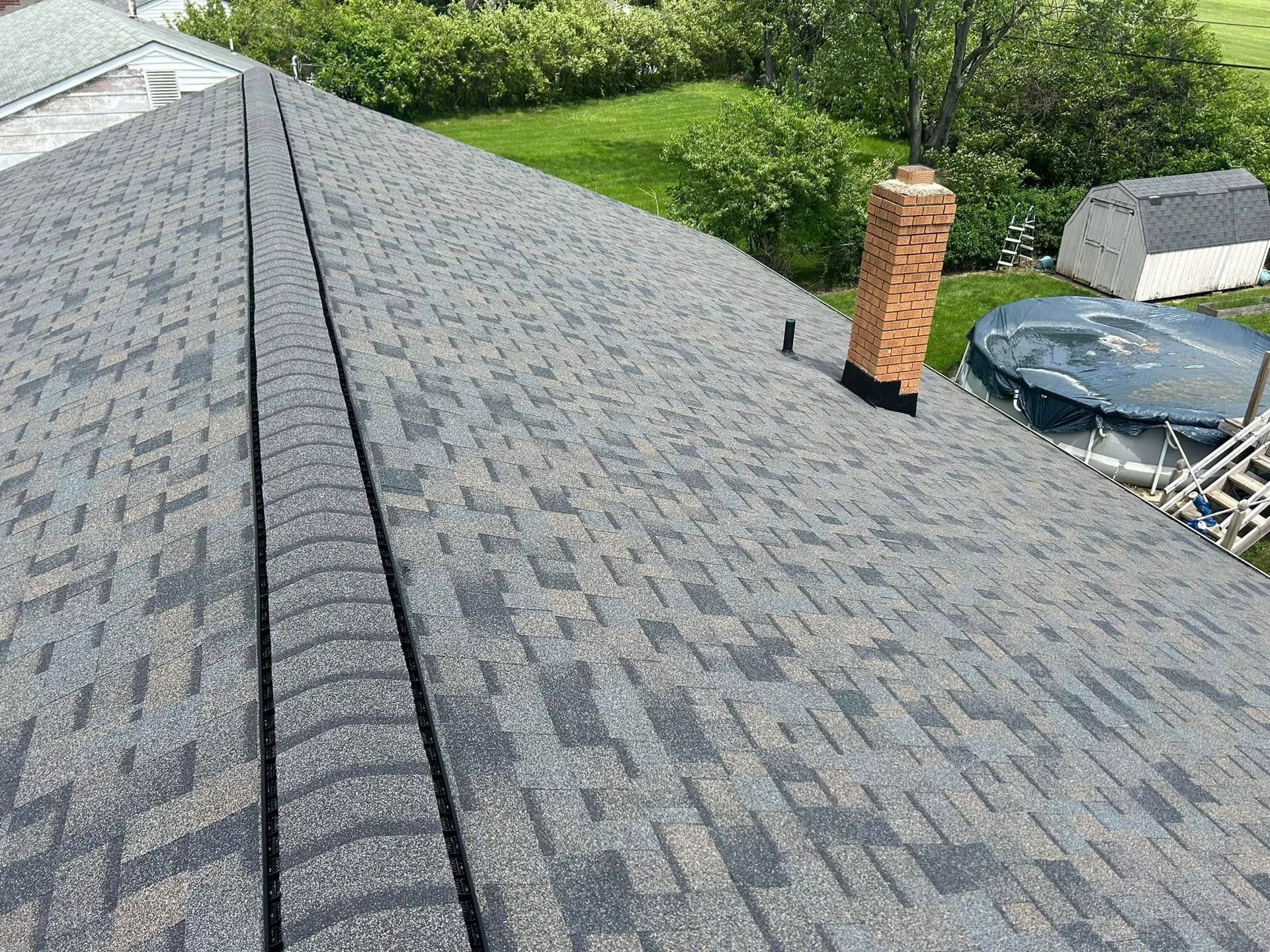 A high-angle view of a gray shingled roof with a brick chimney and a small pipe vent.