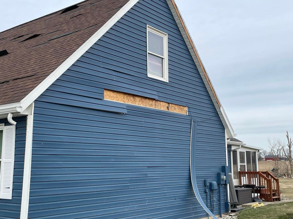 A blue-sided house wall with damaged, missing vinyl siding revealing a strip of exposed wooden sheathing.