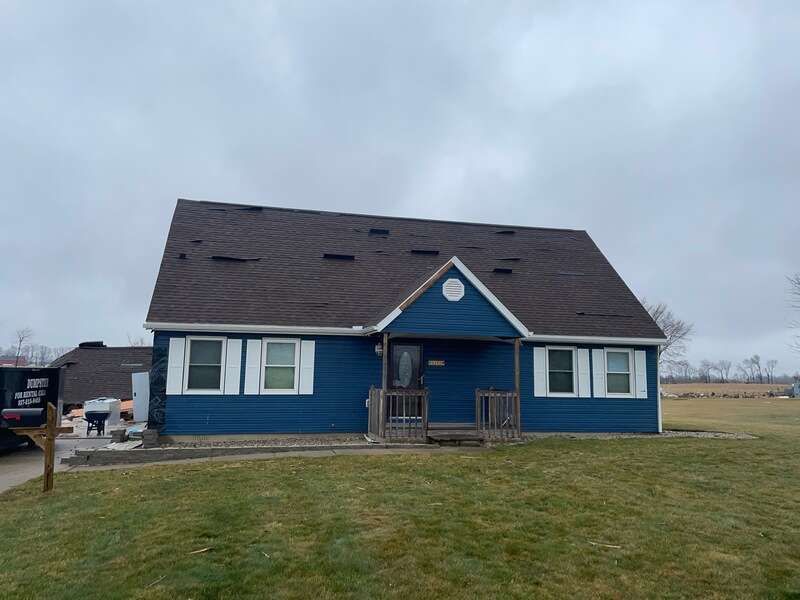 A blue one-story house with white shutters and a brown roof, set in a grassy yard under a cloudy, overcast sky.