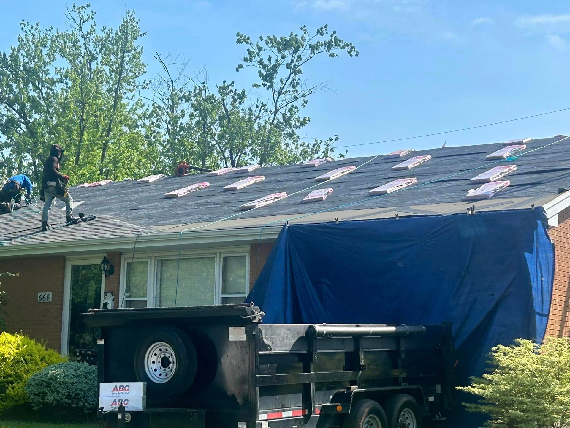 Two workers on a residential roof installing shingles, with a debris trailer parked in the front yard.