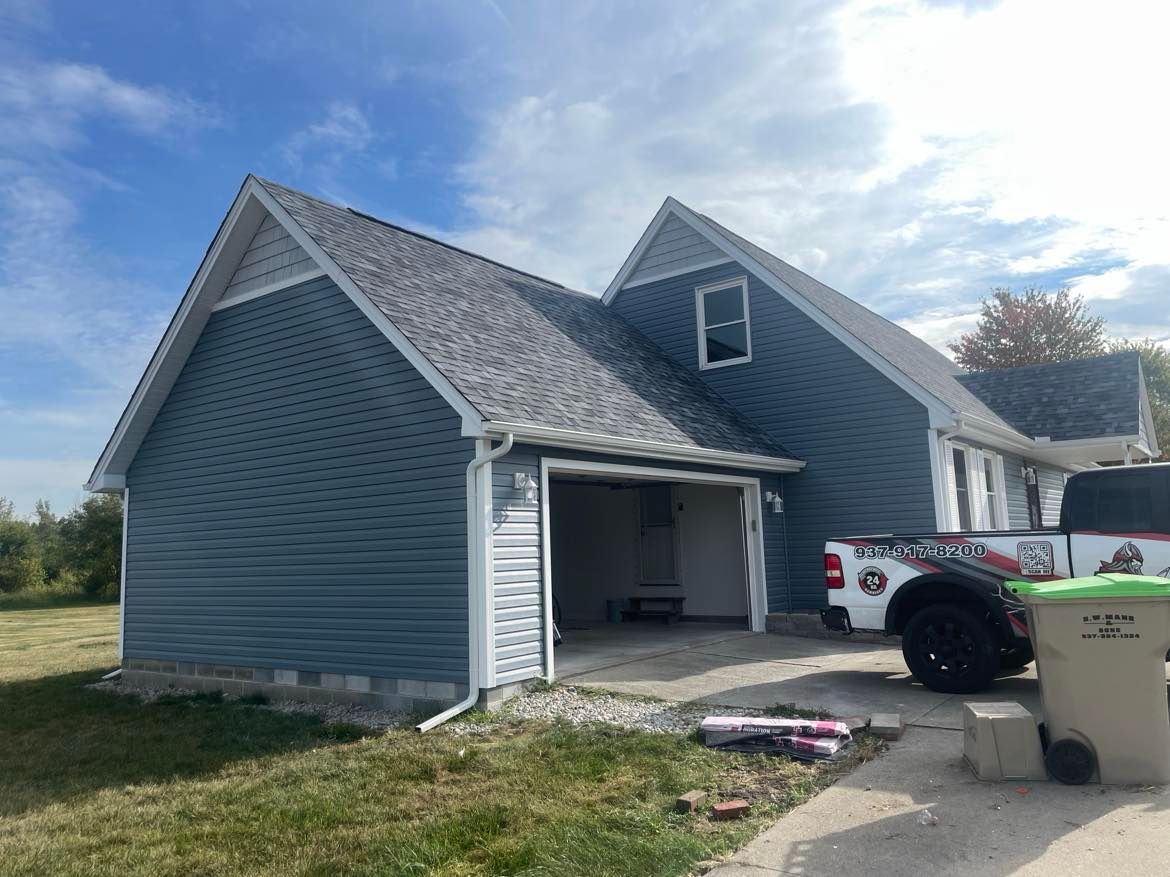 A blue-sided house with a garage and two gabled roofs under a partly cloudy sky, with a truck parked in the driveway.