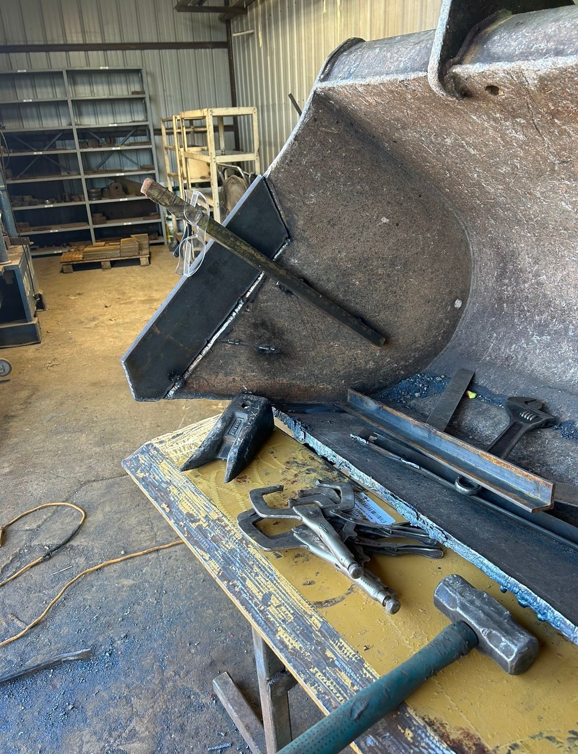 Welding repair on a large metal bucket in a workshop setting with tools and metal pieces on a yellow table.