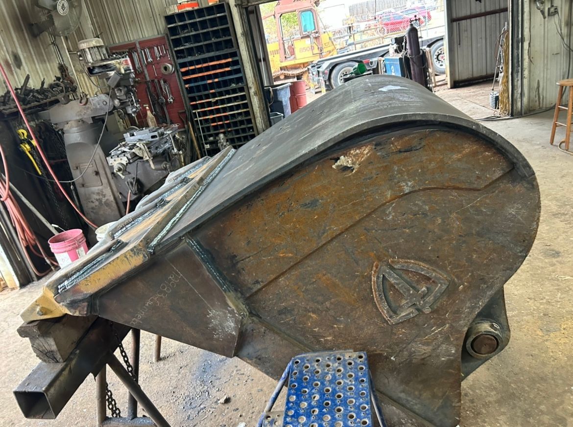 A large, rusty excavator bucket in a workshop. It is supported by a metal frame.