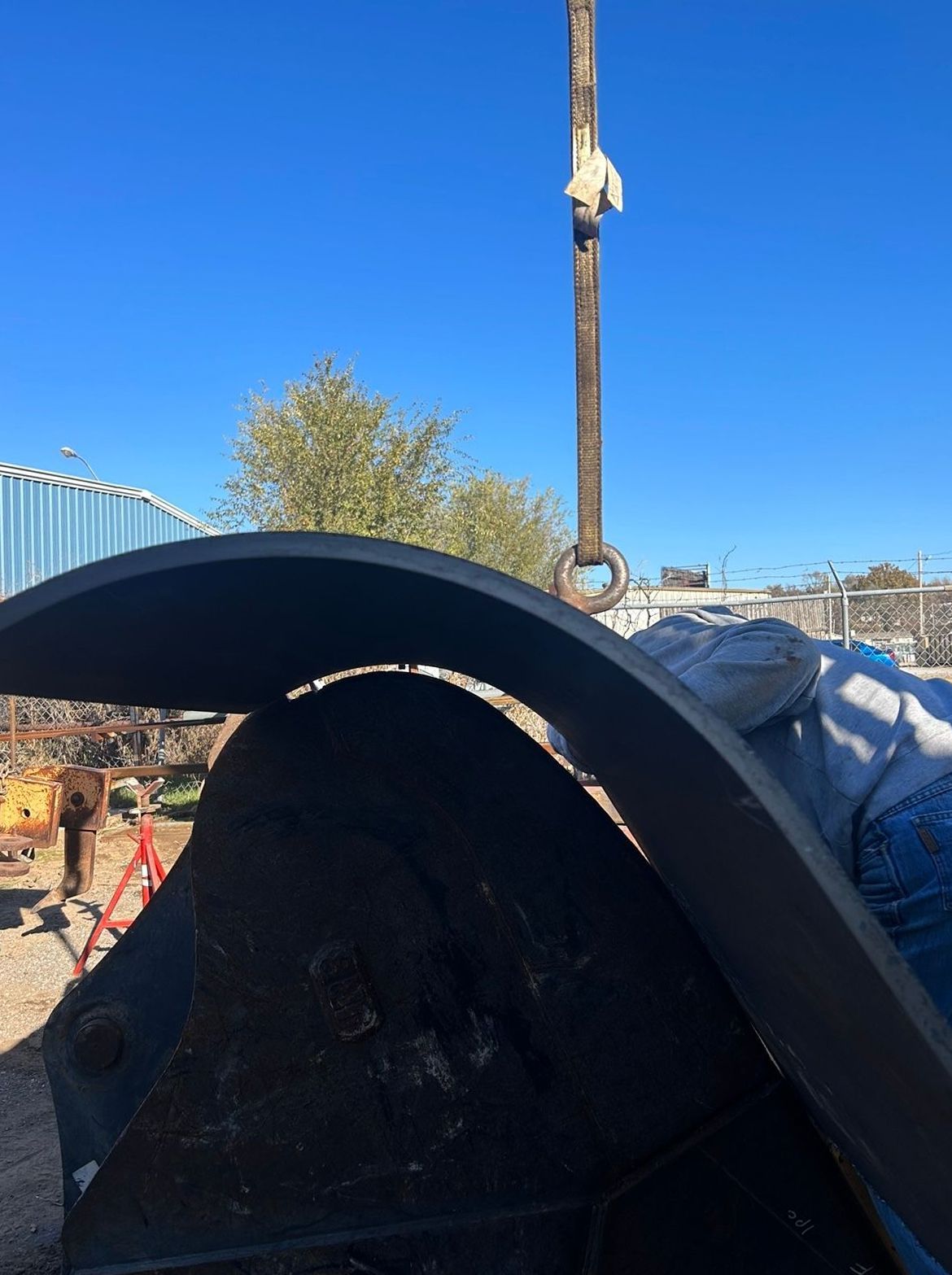 Large metal bucket being lifted by a crane outdoors, with a clear blue sky.
