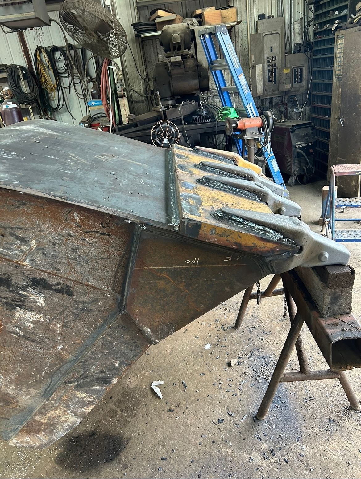 A large, rusty metal bucket being worked on in a workshop; the bucket is propped up on sawhorses.
