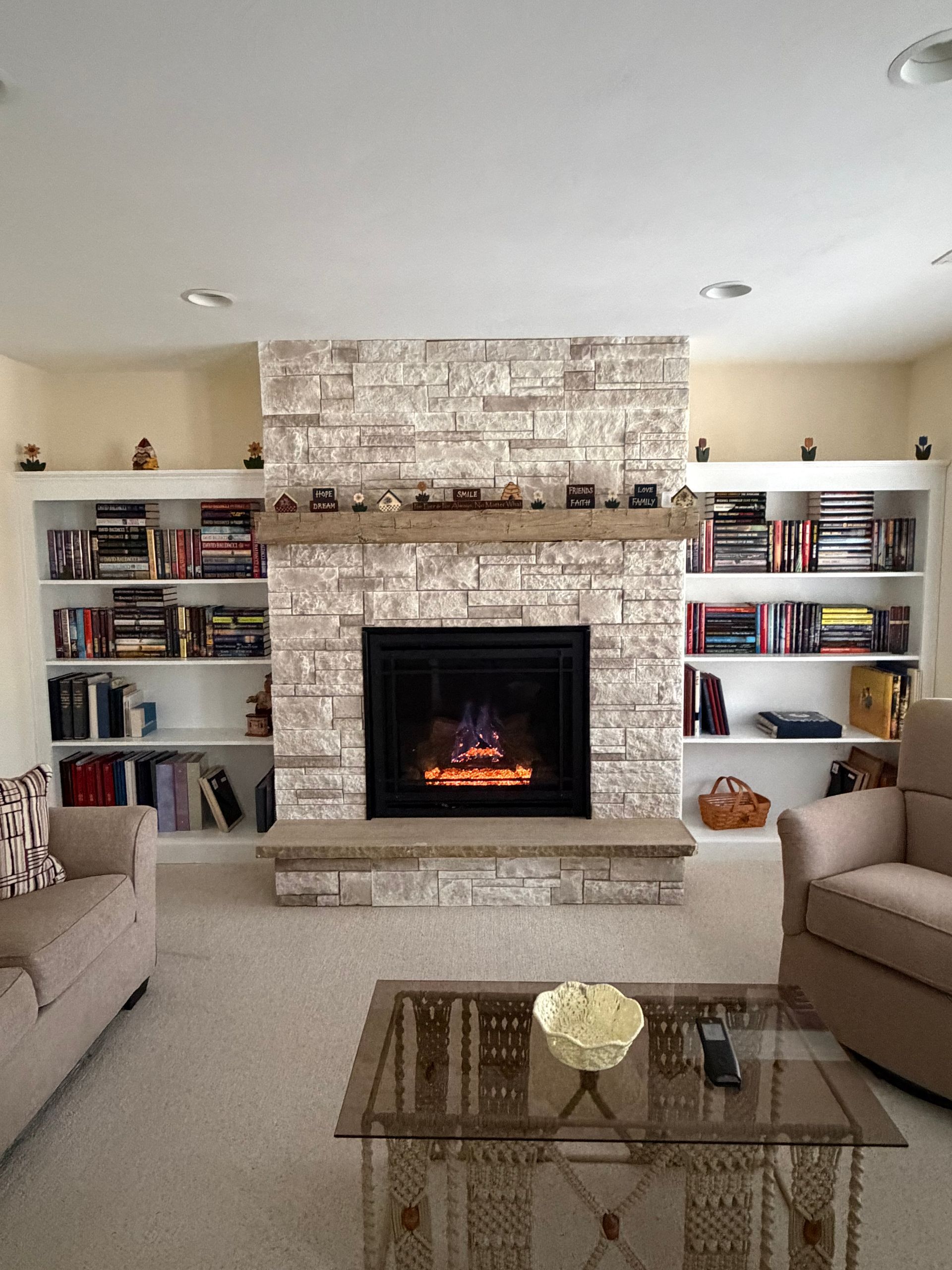 Living room with a stone fireplace, built-in shelves, beige sofas, and a glass coffee table