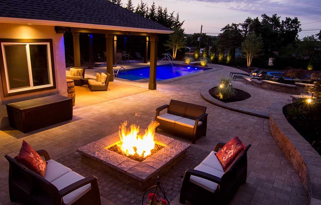 Cozy backyard patio at dusk with a lit fire pit, cushioned chairs, and warm landscape lighting