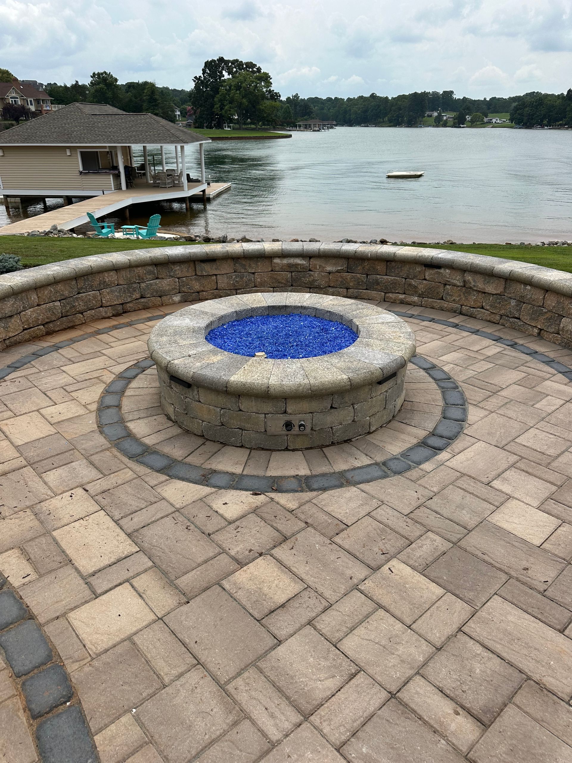 Stone fire pit with blue glass centered on a brick patio by a lake with trees and small docks in the background