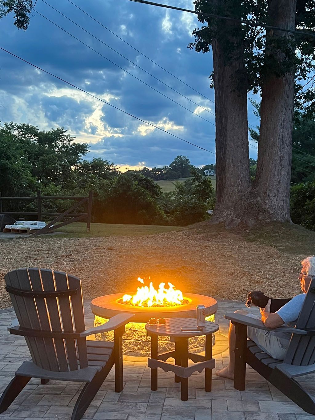 Person sitting by a backyard fire pit at dusk, with trees and cloudy sky in the background