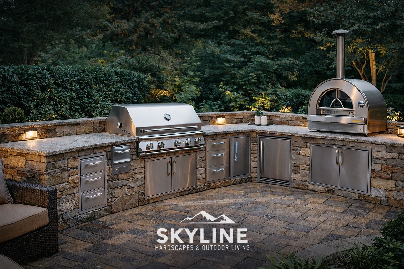 Outdoor stone patio with built-in stainless grill, pizza oven, and cabinets under string lights, surrounded by greenery