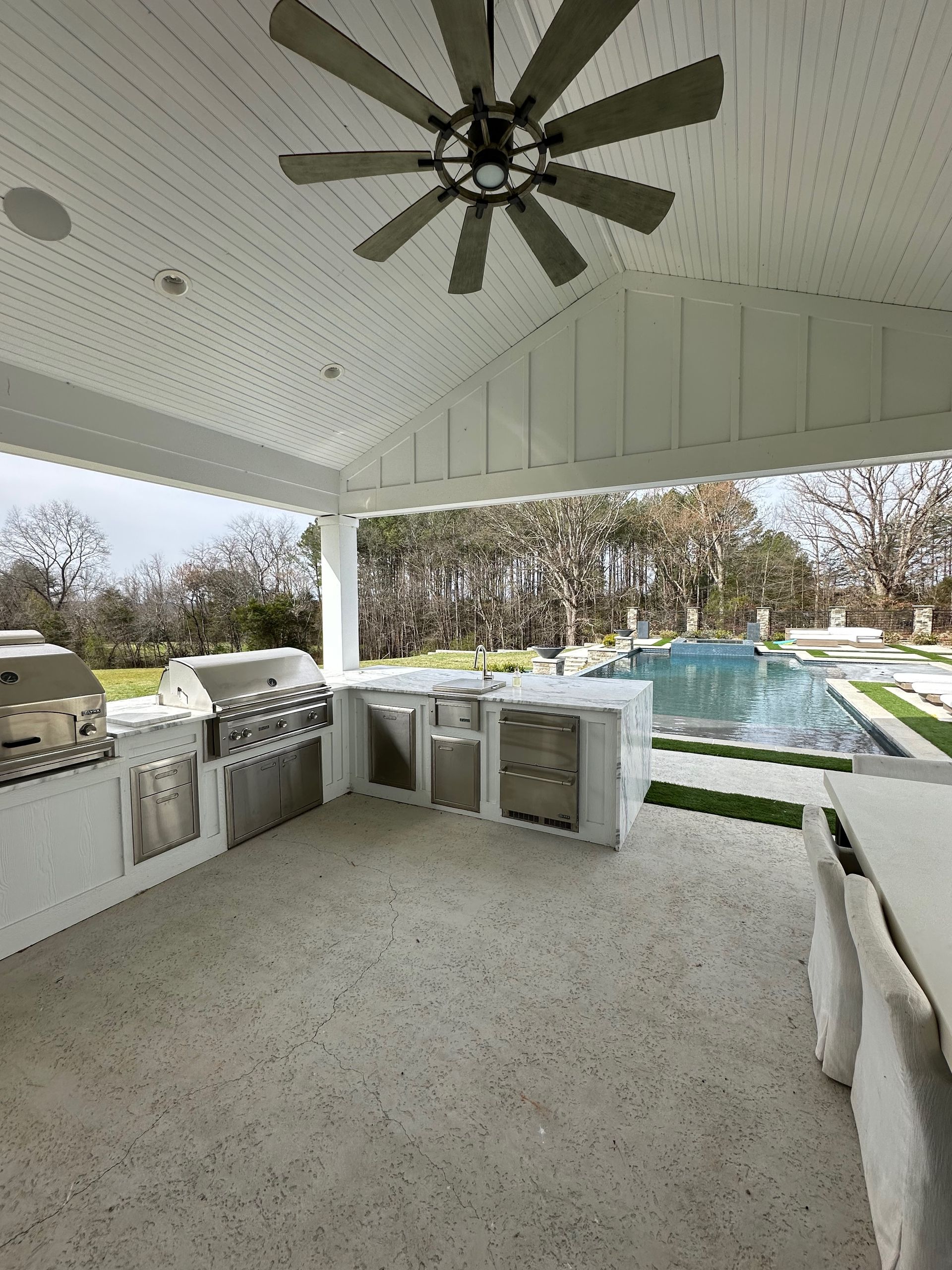 Covered outdoor patio with ceiling fan, grill, and bar seating overlooking a pool and yard.