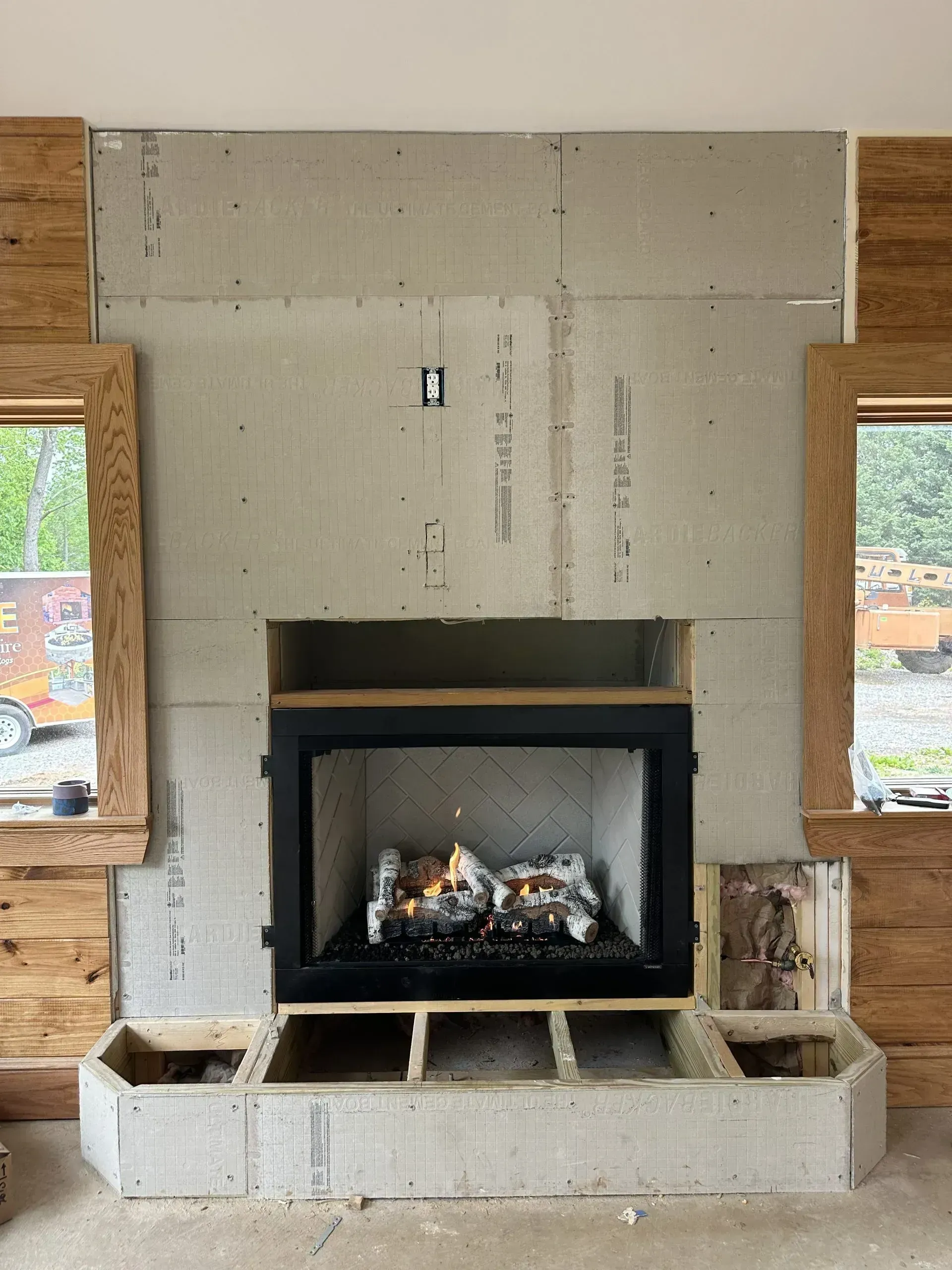 Fireplace under construction, black insert with gas logs, surrounded by light-colored stonework and wood accents.