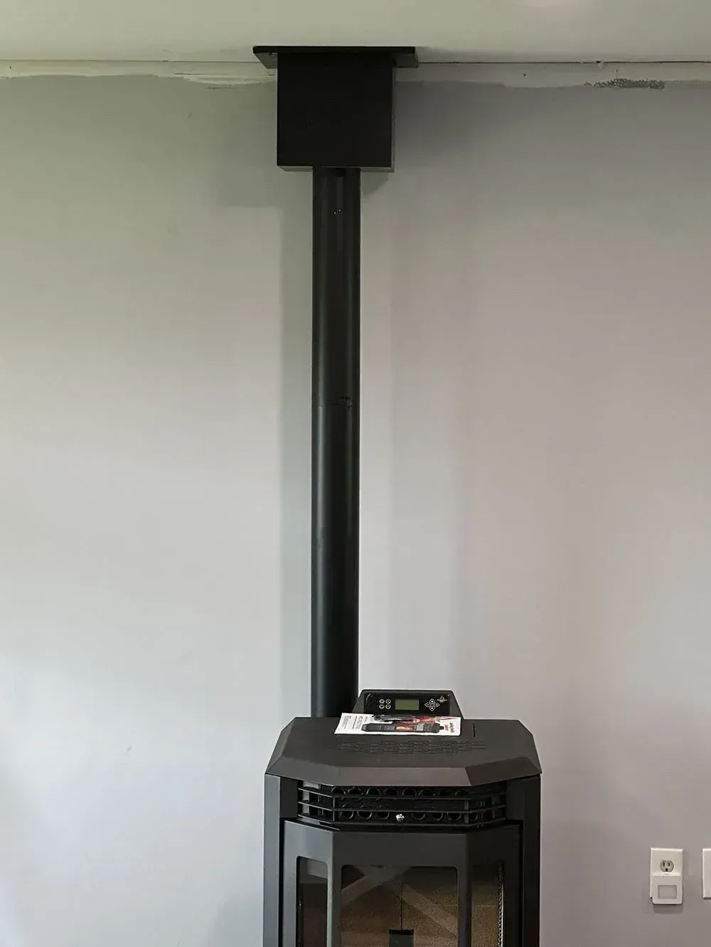 Black stove with tall black pipe against a gray wall, reaching the ceiling.