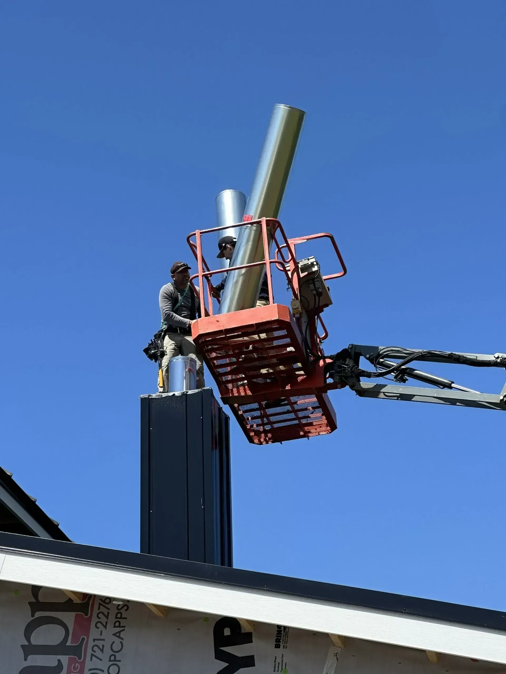 Construction workers installing a metal chimney pipe using a lift on a rooftop, against a blue sky.