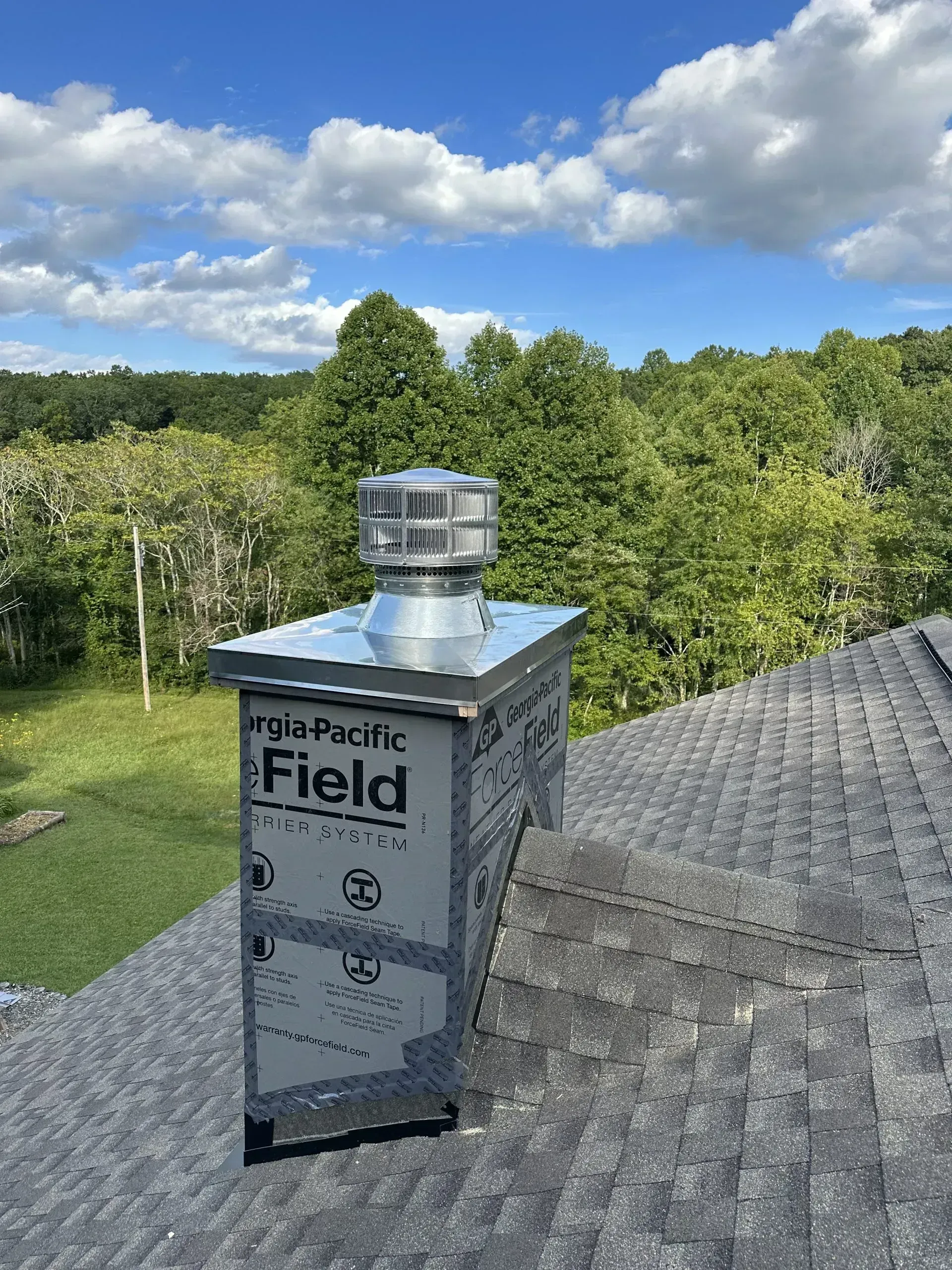 Chimney on a roof with a metal cap and cap, blue sky and trees in background.