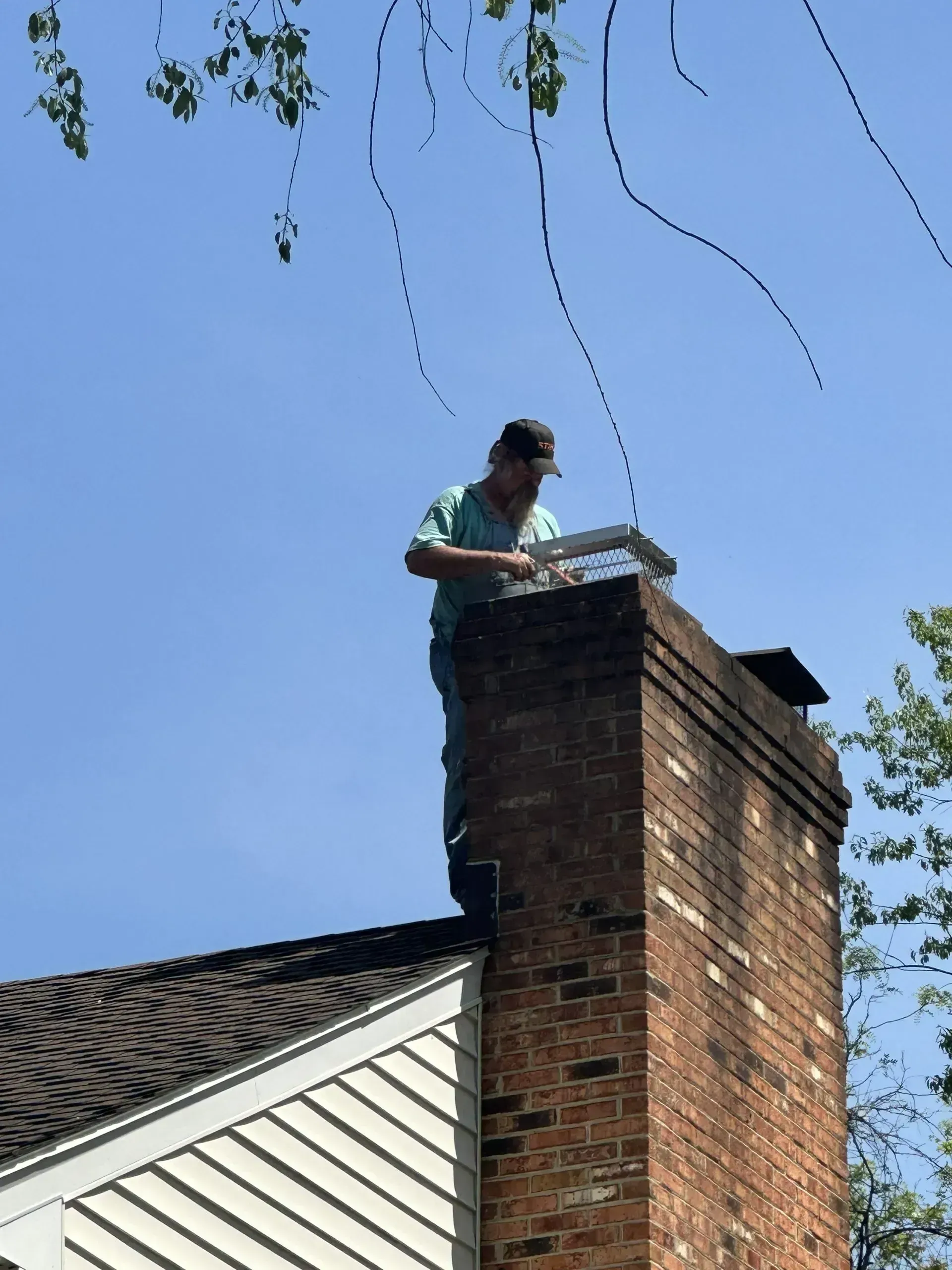 Person on a chimney, working on a square object against a blue sky, branches above.