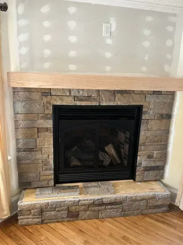 Fireplace with stone facade, wood mantel, and unfinished drywall above.