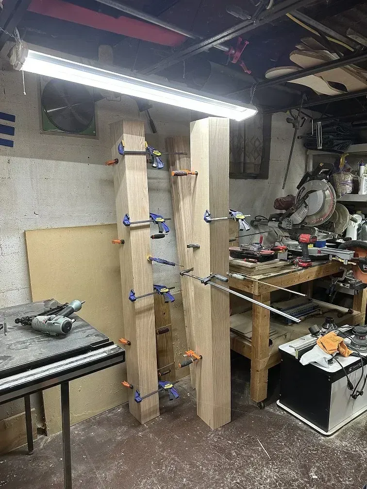Wooden beams clamped together in a workshop under fluorescent lighting. A table saw and tools are nearby.