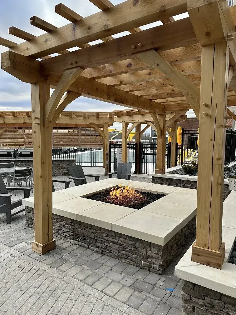 Pergola over a stone fire pit, with seating and a pool area in the background.
