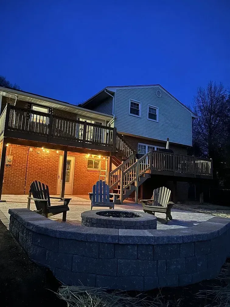 Backyard patio with fire pit, Adirondack chairs, and a house with a deck at dusk.