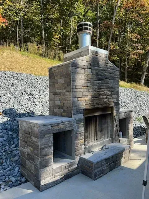 Stone outdoor fireplace with chimney, built-in storage, and bench. Gray stone against a backdrop of foliage and gravel.