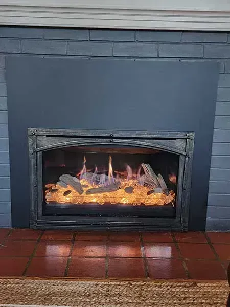 Gas fireplace with flames, framed by a dark grey surround on a brick hearth.
