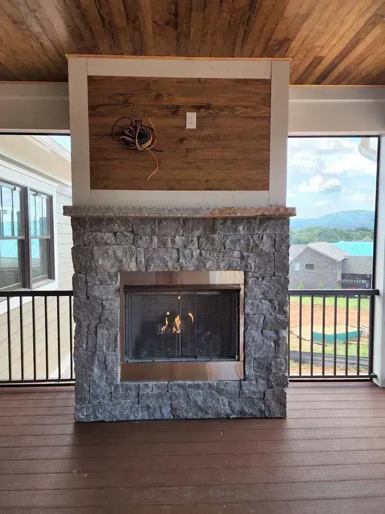 Stone fireplace on a deck, with wood surround, mounted TV area, flames visible, and outdoor view.