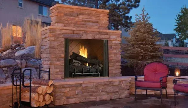 Outdoor stone fireplace with a lit fire, beside red chairs and a stack of firewood.