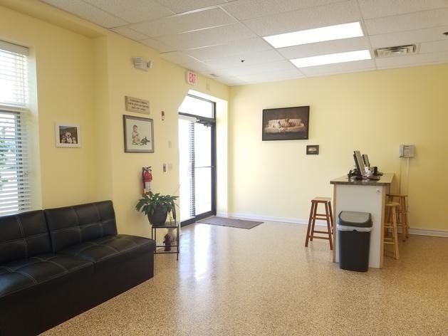 A light yellow office reception area with a black sofa, a small check-in desk, a trash can, and a glass-paneled entry door.