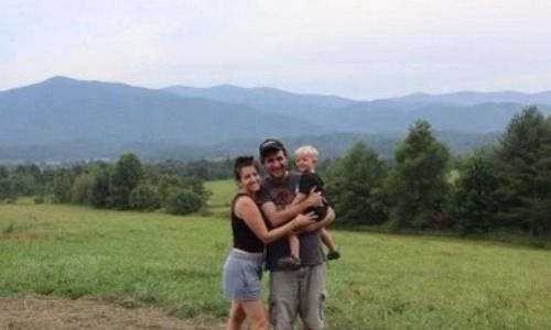 A family stands together, embracing in a grassy field with rolling mountains in the distance under a cloudy sky.
