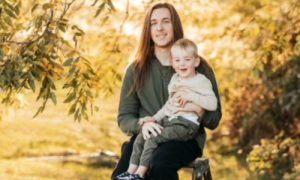 A long-haired person sitting outdoors in a green shirt, holding a young child on their lap. Soft autumn lighting.