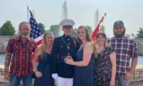 A group of six people posing for a photo outdoors in front of a fountain and American flags, with a service member centered.