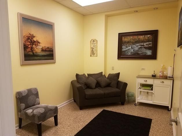 A waiting room with yellow walls, a small gray couch, a patterned accent chair, a black rug, and two landscape paintings.
