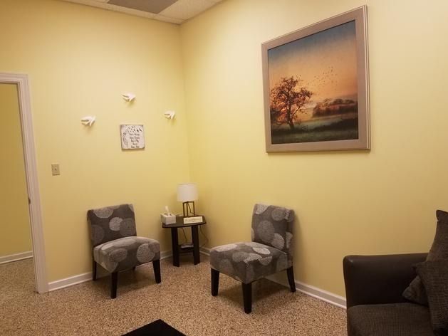 A waiting room with yellow walls, two gray patterned chairs, a small table with a lamp, and a landscape painting.