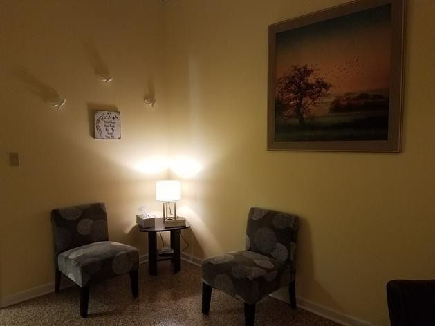 Two gray patterned chairs sit in a cozy, softly lit corner next to a small side table with a lamp and a framed landscape.