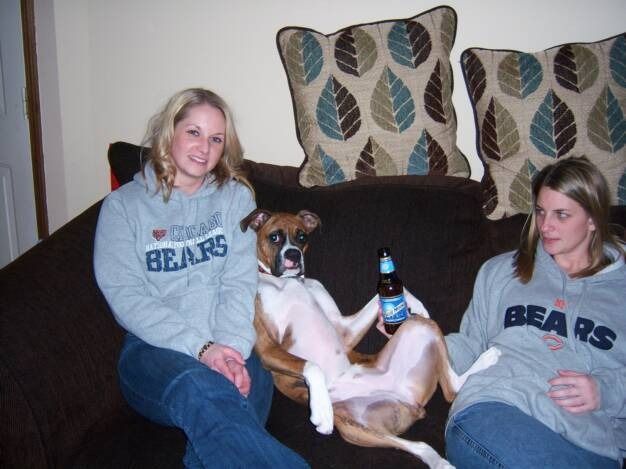 Two women in grey Bears hoodies sit on a sofa with a dog between them that is sitting upright and holding a beer bottle.