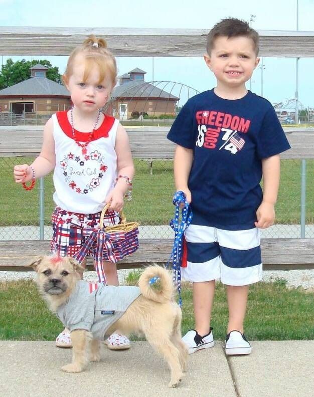 Two children standing outdoors next to a small, tan, short-haired dog wearing a gray shirt, with a wooden fence behind them.