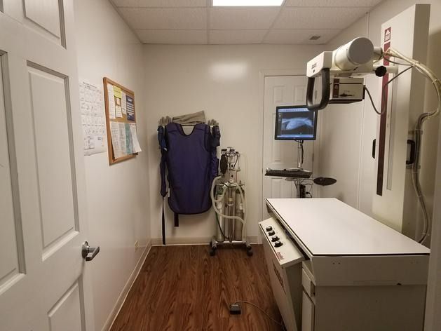 Small room containing an X-ray imaging machine, a protective lead apron hanging on the wall, and a computer monitor.