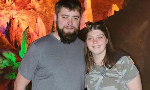 A person with a beard and a person with long hair smiling together in front of illuminated cave rock formations.