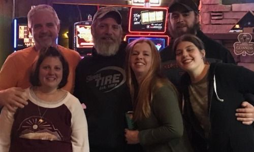 A group of six smiling people posing for a photo inside a bar with neon gaming machines in the background.