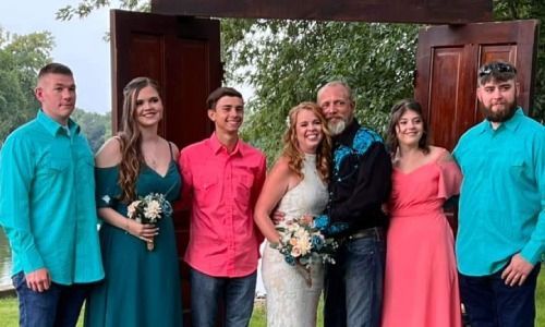 A wedding party of seven stands outdoors in front of a rustic wooden door frame, posing for a group portrait.