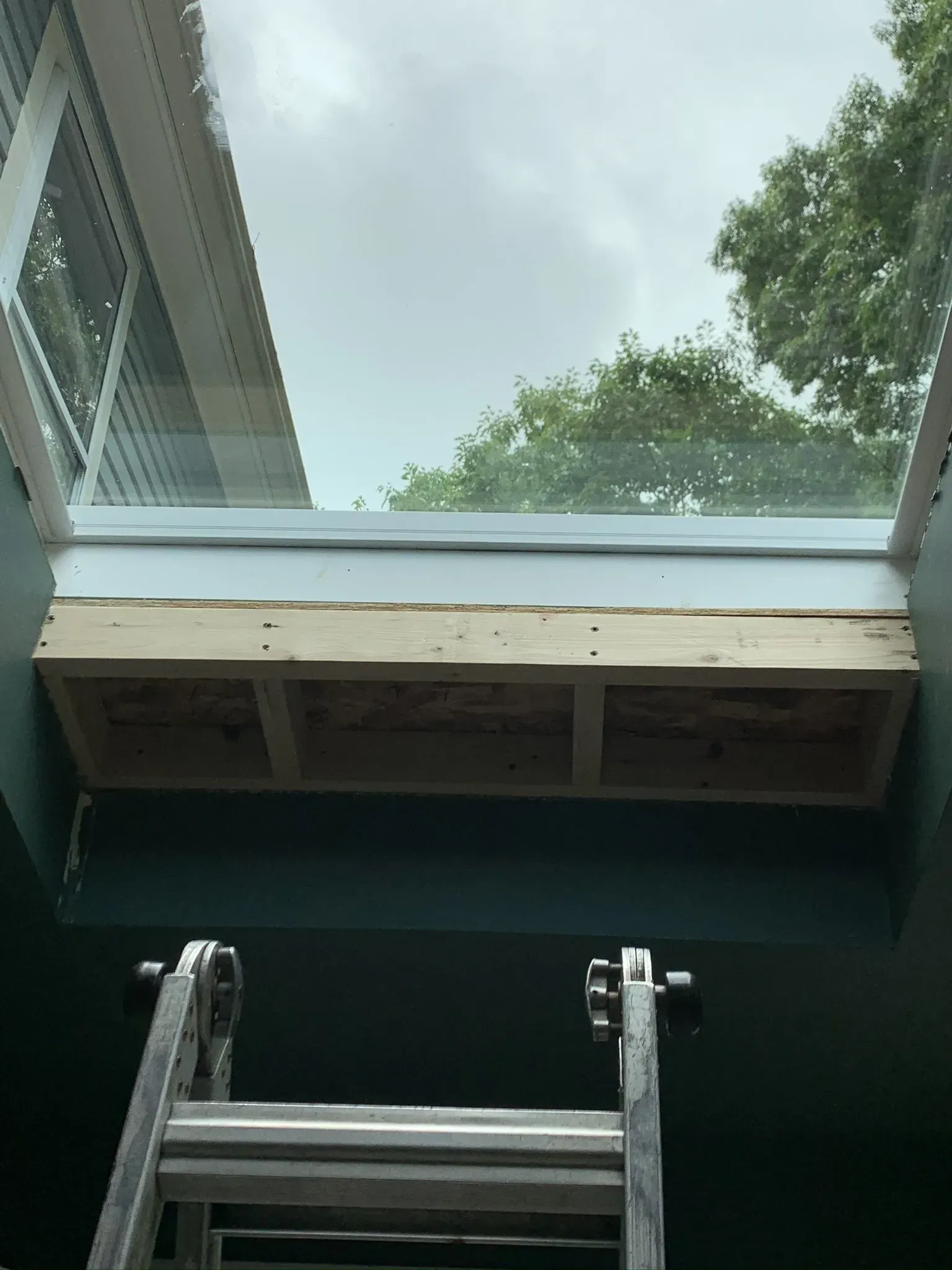 Looking up at a skylight with a wooden frame installed, ladder in the foreground, cloudy sky visible.