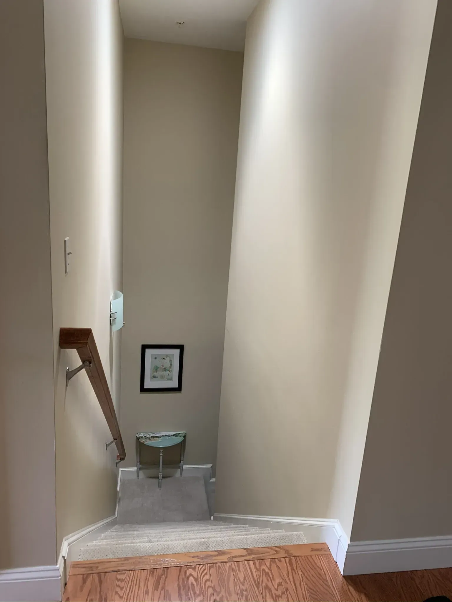 Stairwell with beige walls, wooden handrail, and carpeted landing. Small table and framed art at the bottom.