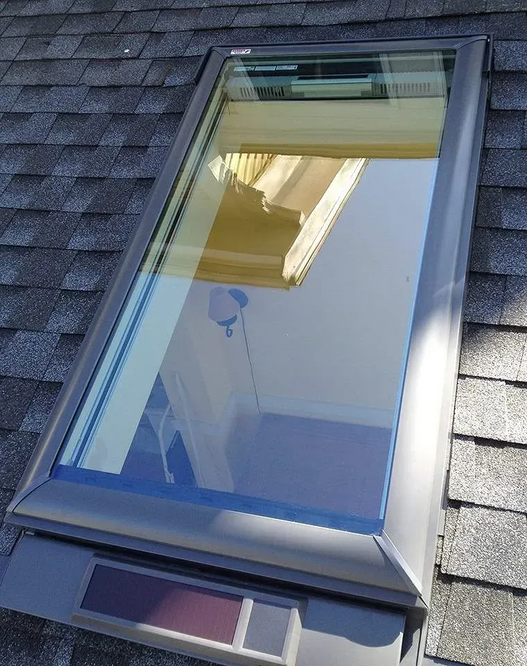 Skylight on a shingle roof reflects a yellow building and a room with a blue wall.